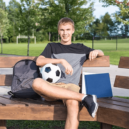 Jeune &eacute;tudiant assis sur un banc dans un parc avec un ballon de football et des cahiers scolaires, illustrant l&rsquo;&eacute;quilibre entre sport et &eacute;ducation