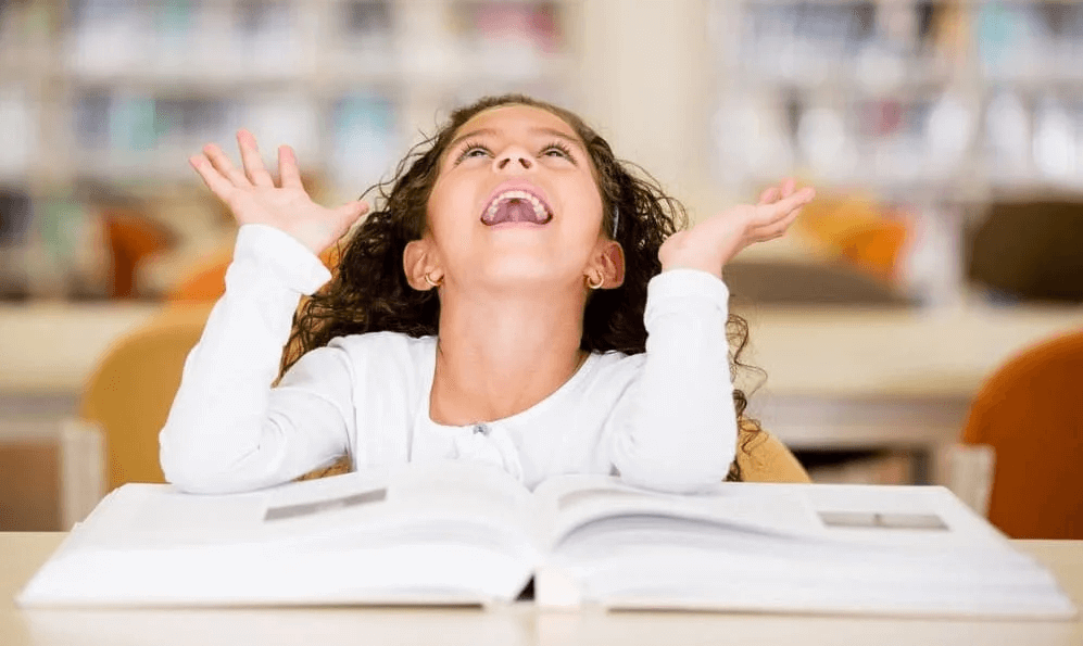 Petite fille joyeuse levant les mains en lisant un livre ouvert à la bibliothèque