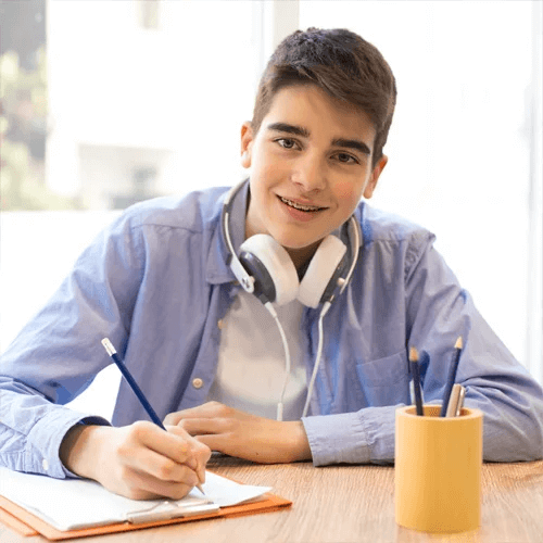 Adolescent souriant avec casque audio autour du cou, &eacute;crivant dans un cahier pendant une s&eacute;ance d&rsquo;&eacute;tude &agrave; domicile