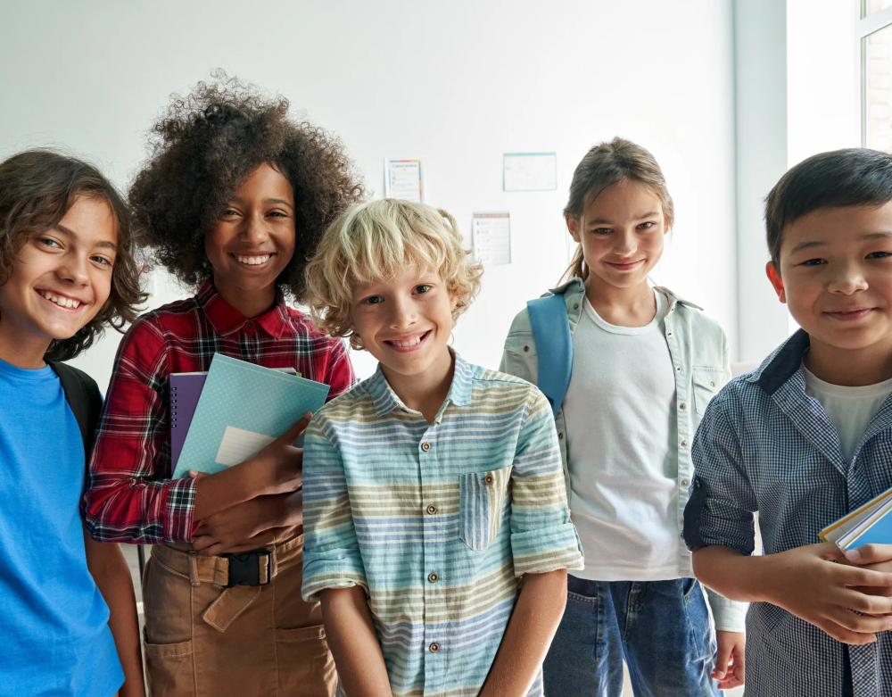 Cinq enfants heureux posant ensemble dans un couloir d’école, portant des cartables et des cahiers, symbolisant l’éducation et la diversité