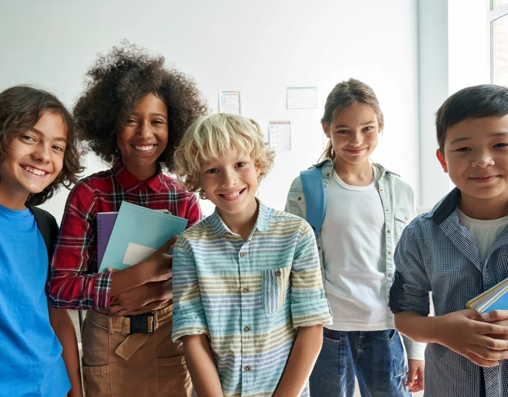 Cinq enfants heureux posant ensemble dans un couloir d’école, portant des cartables et des cahiers, symbolisant l’éducation et la diversité