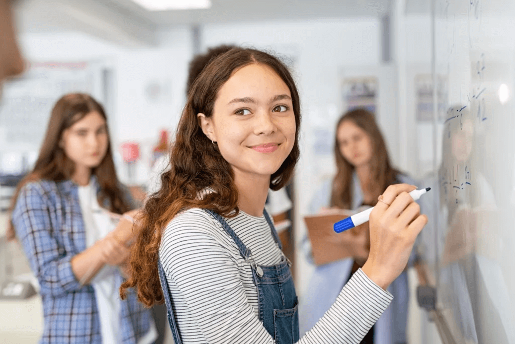 Jeune fille avec un marqueur bleu écrivant sur un tableau blanc, entourée de camarades de classe