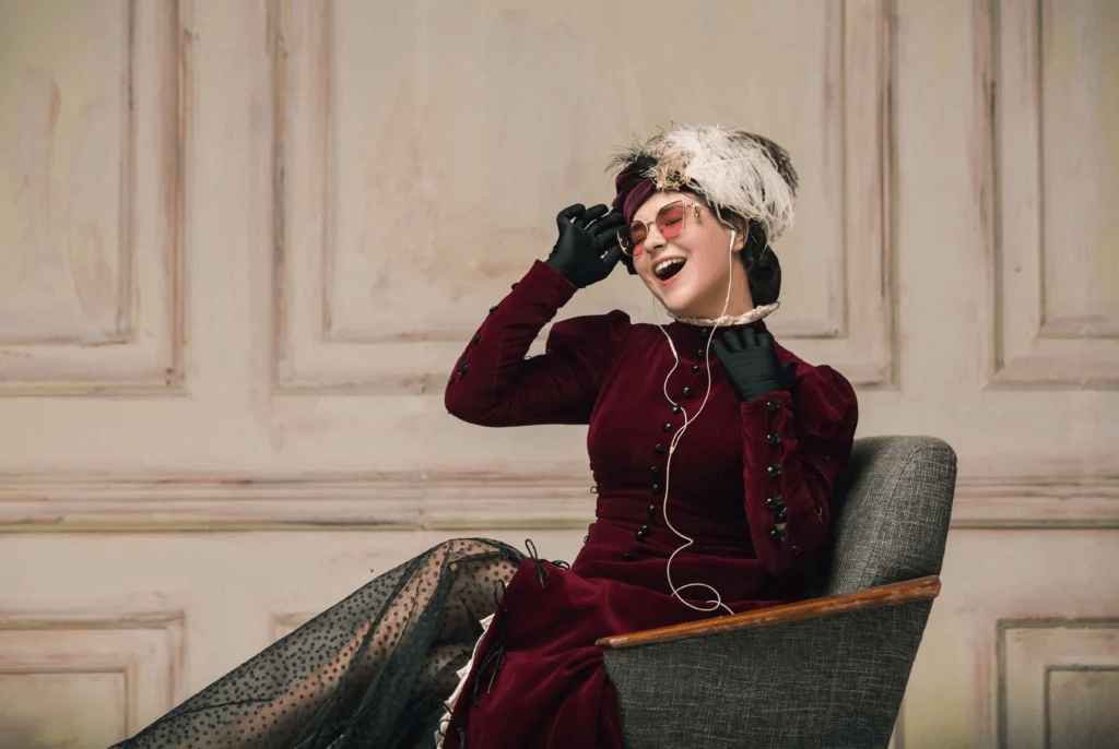Portrait d’une femme en robe vintage bordeaux avec chapeau à plumes, lunettes rétro et écouteurs, assise sur une chaise et profitant de la musique
