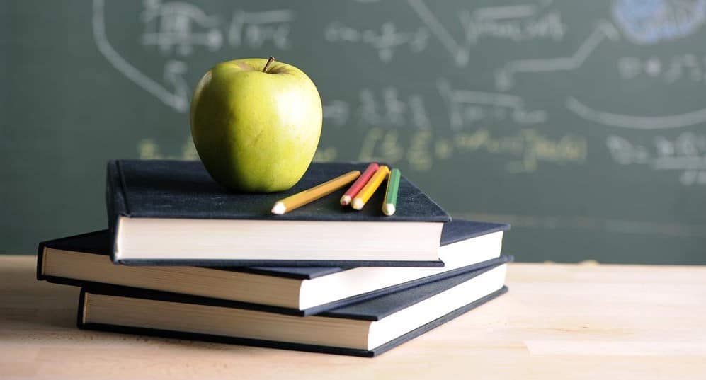 Pomme verte posée sur une pile de livres scolaires avec des crayons de couleur, tableau noir en arrière-plan.