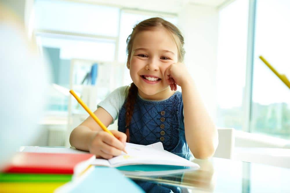 Fillette souriante écrivant au crayon dans un cahier, assise à une table de classe lumineuse
